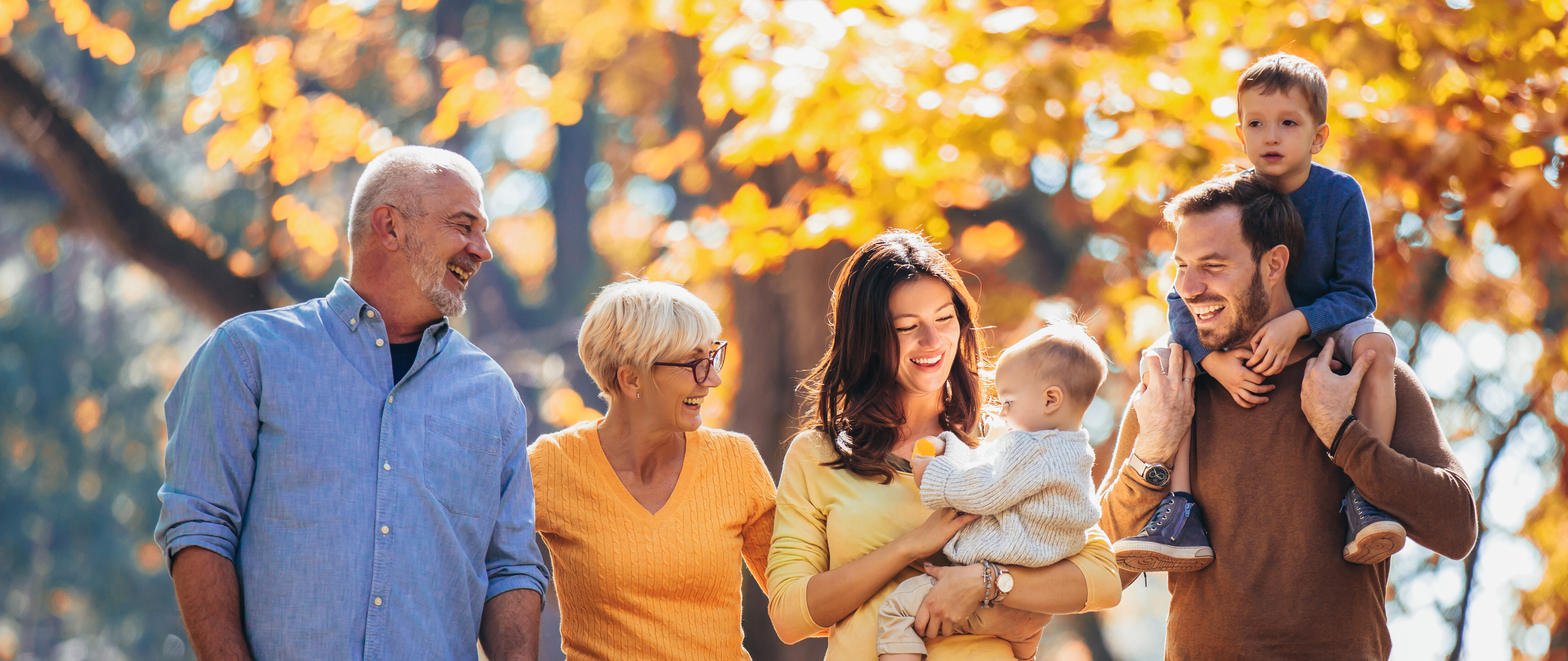 Eine Gruppe von Menschen macht einen Herbstspaziergang: ein älterer Mann, eine ältere Frau, eine junge Frau mit kleinem Kind auf dem Arm und ein Mann mit einem kleinem Jungen auf seinen Schultern.