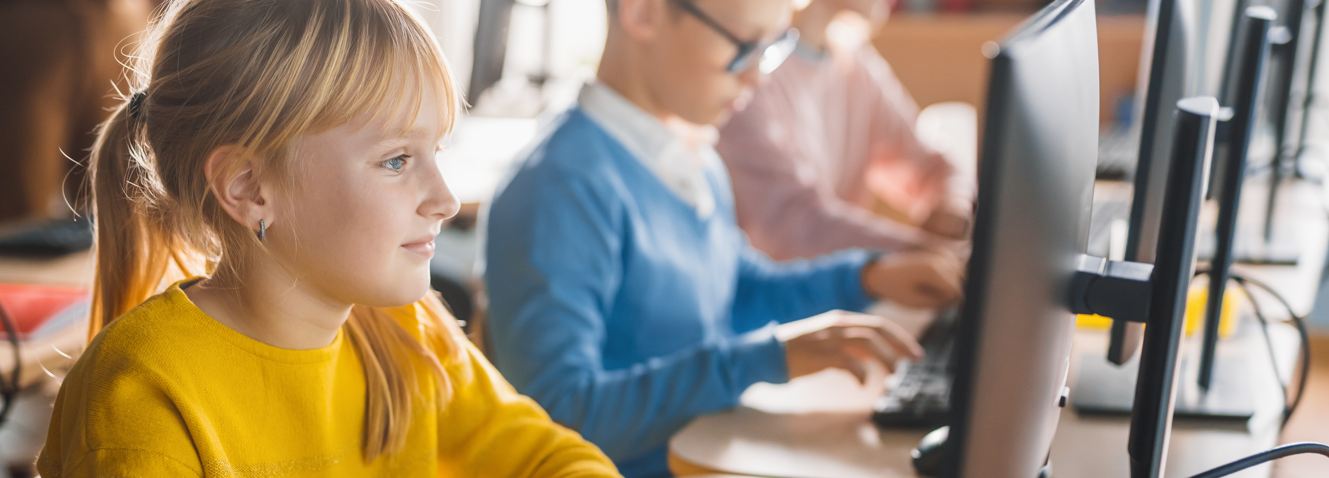 Drei Personen sitzen nebeneinander auf Bürostühlen an Computern an Schreibtischen. Eine Frau tippt auf einer Tastatur und hält eine Computermaus in ihrer Hand.