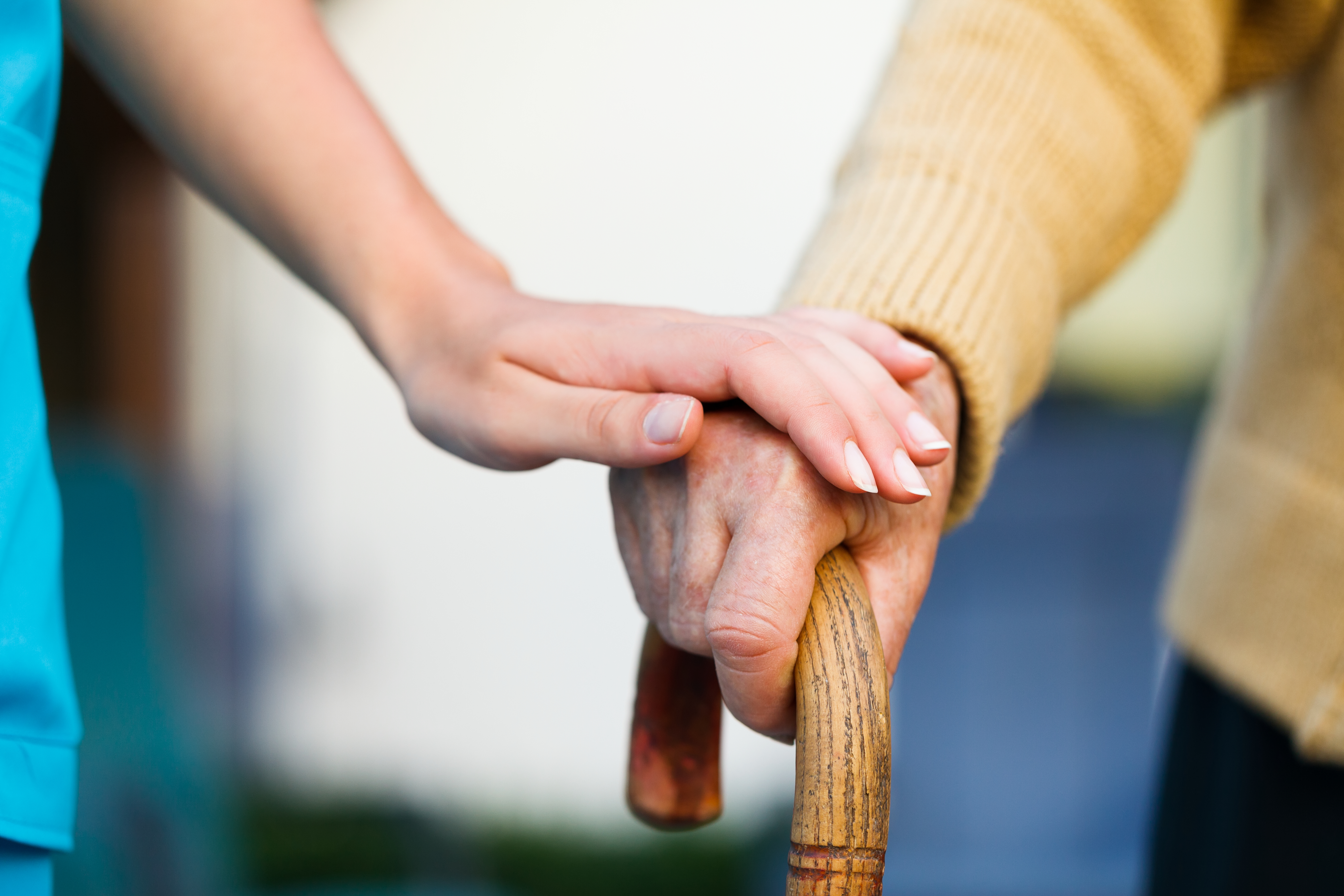 Eine junge Hand auf der Hand einer alten Person, die sich auf einen Gehstock stützt.