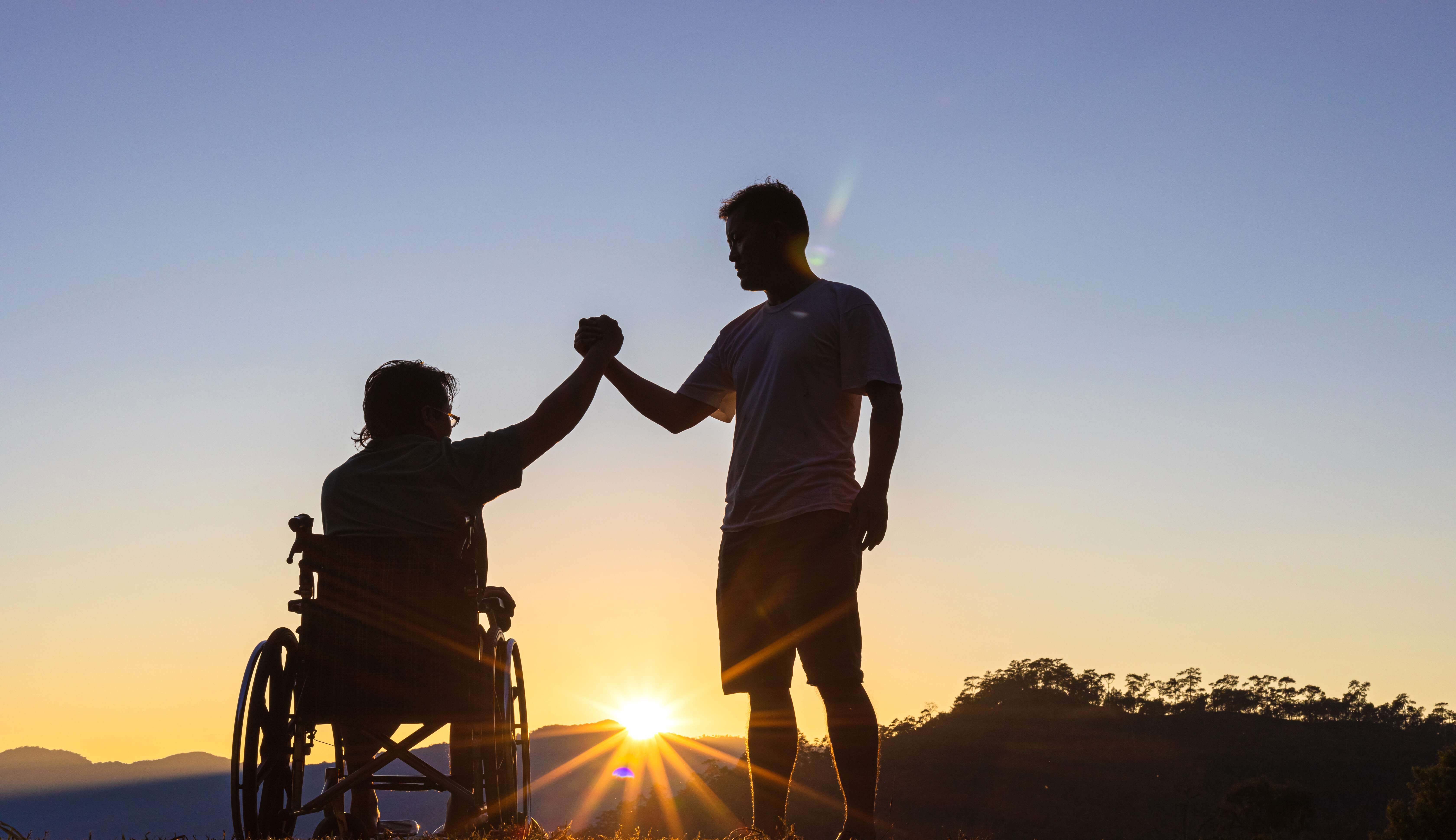 In Siegerpose reichen sich ein Mann im Rollstuhl und ein anderer Mann die Hände. Sie sind auf einem Hügel vor der untergehenden Sonne.