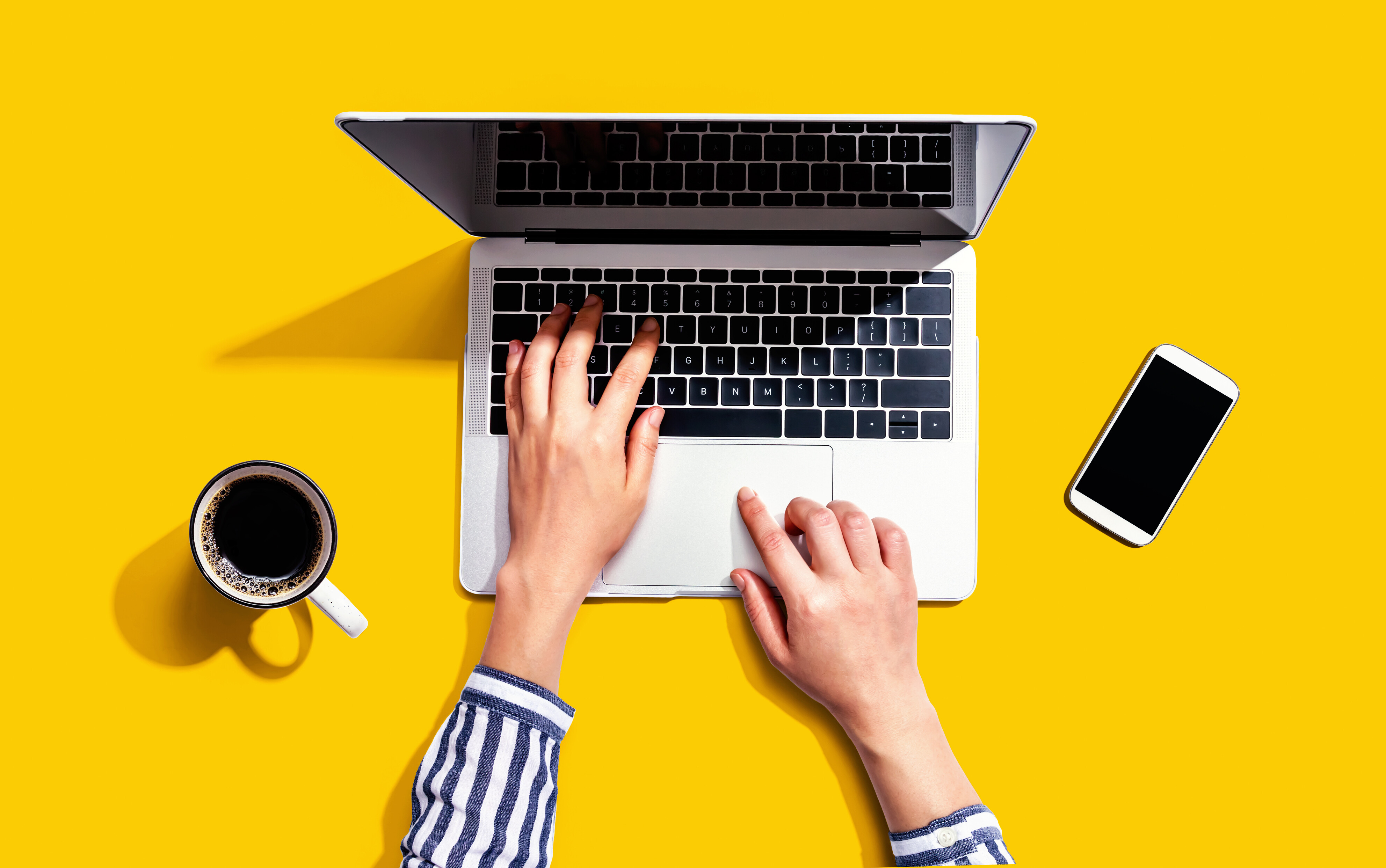 Auf einer gelben Fläche stehen ein Computer und eine Tasse Kaffee, daneben liegt ein Handy. Zu sehen sind die Hände einer Person, die an dem Computer arbeitet.