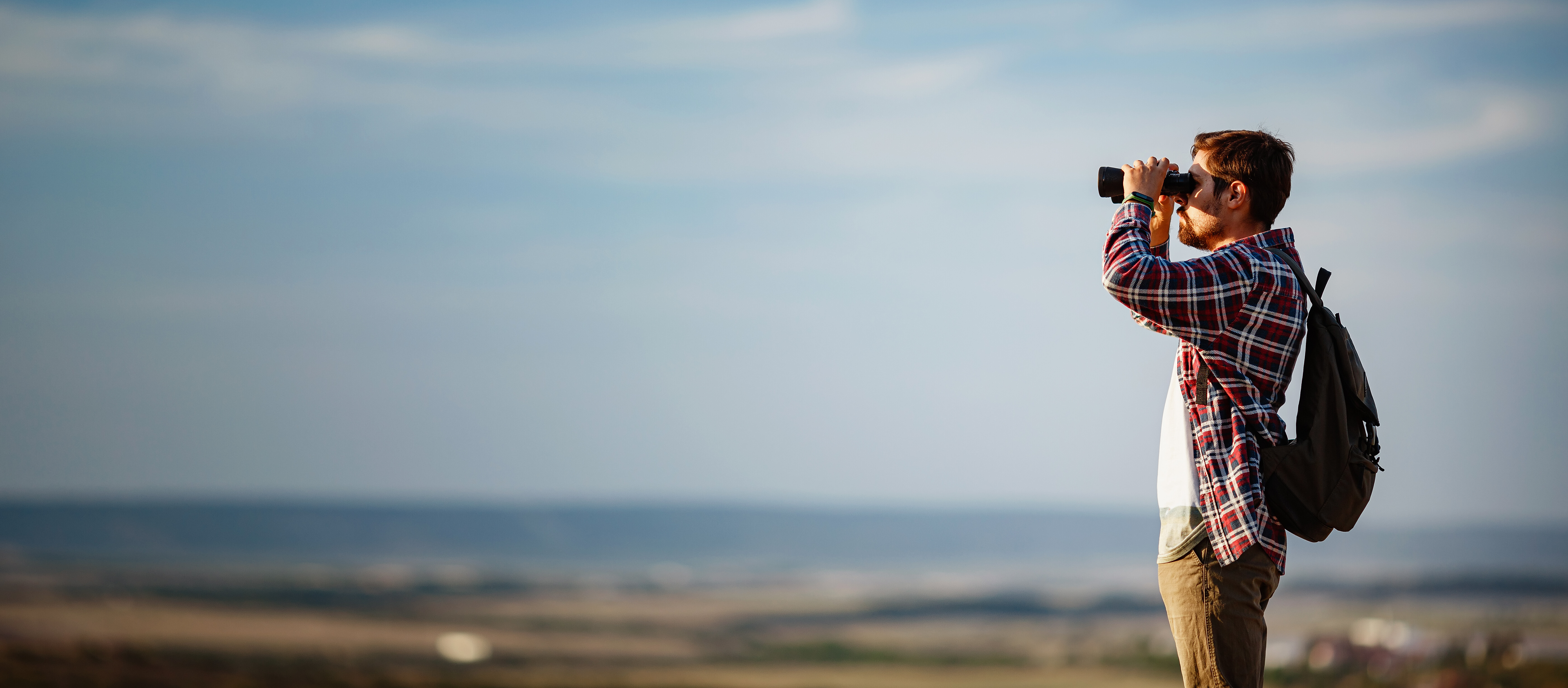 Ein Mann steht auf einem Hügel, trägt einen Rucksack und schaut mit einem Fernglas in die Landschaft.
